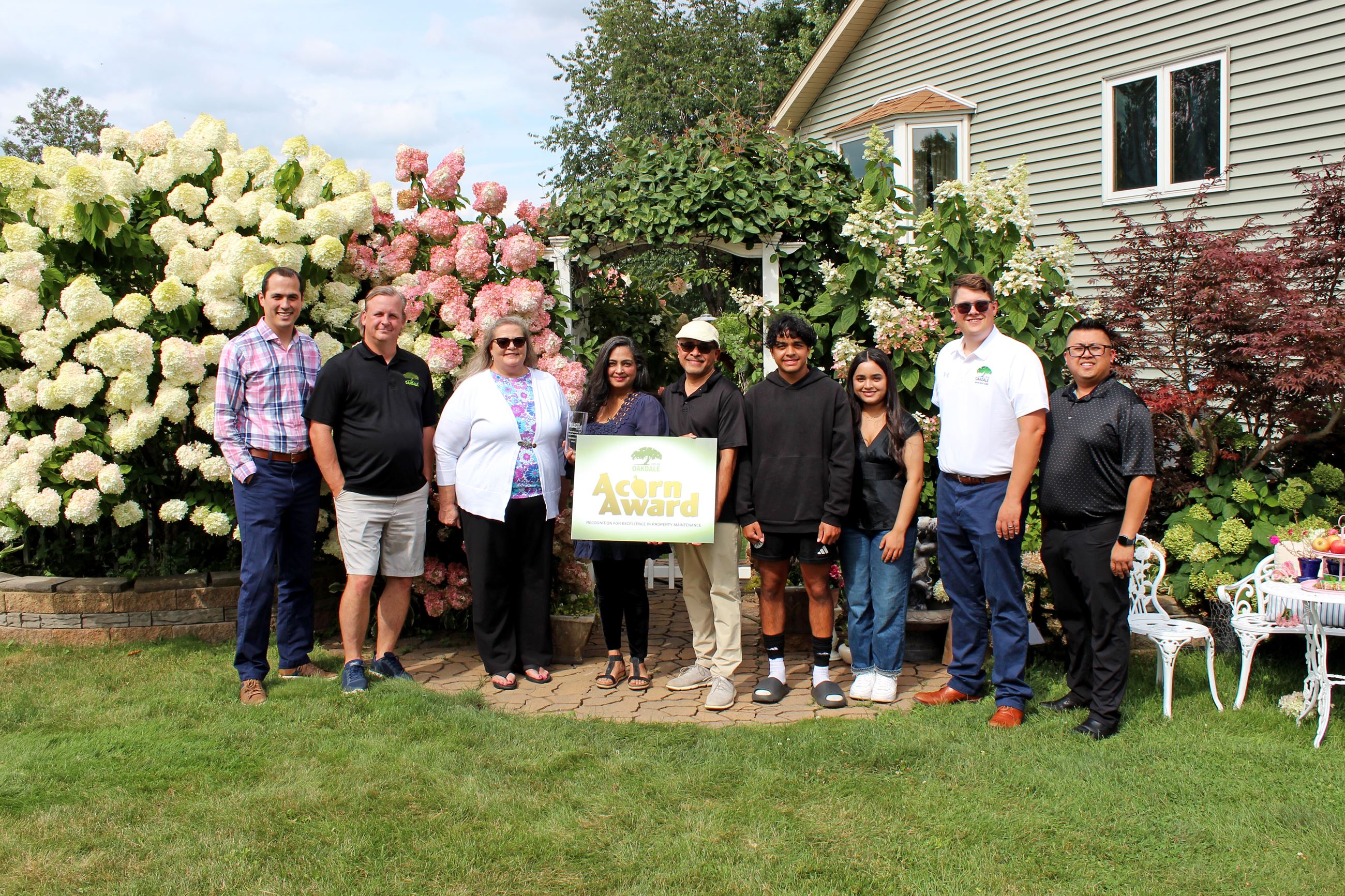 Group of nine people holding a sign that reads "Acorn Award" pose in a garden with white gazeb